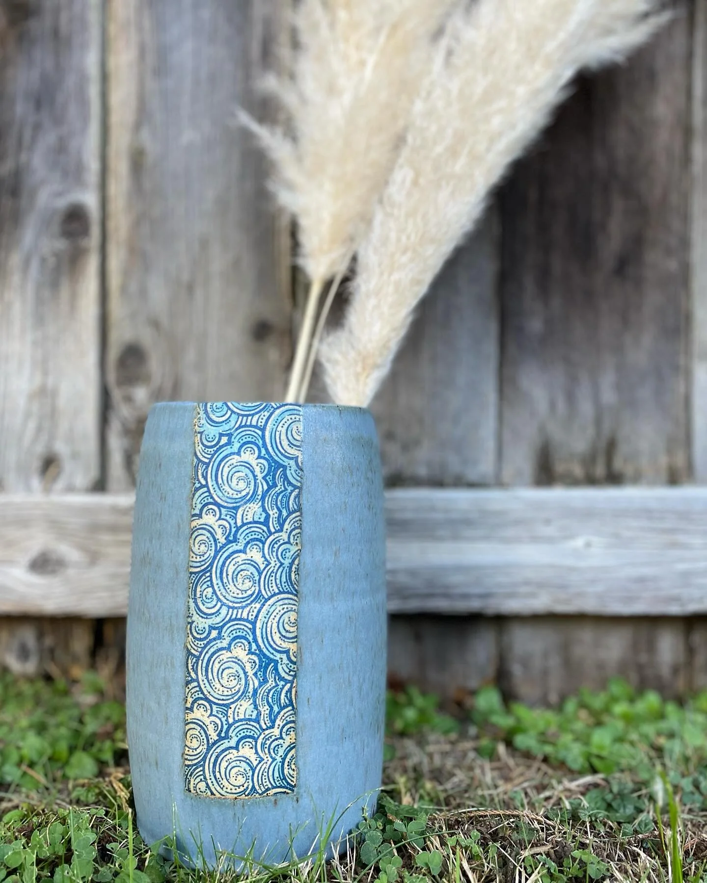 Blue decorative vase with swirl pattern, containing pampas grass, placed on grass near a wooden fence. Sonia Lesage Ceramics