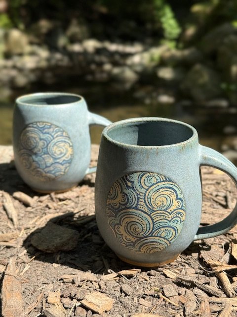 Two ceramic mugs with a stormy skies design, on a log near a river or stream, with blurred green foliage in the background. Sonia Lesage Ceramics