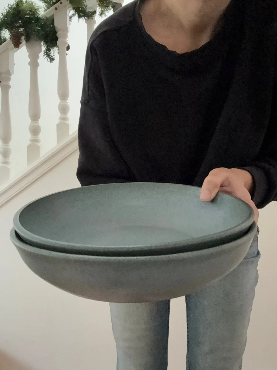 Person holding large blue/grey platters in front of their body, wearing a black sweater and blue jeans, standing indoors near a staircase with holiday decorations. Sonia Lesage Ceramics