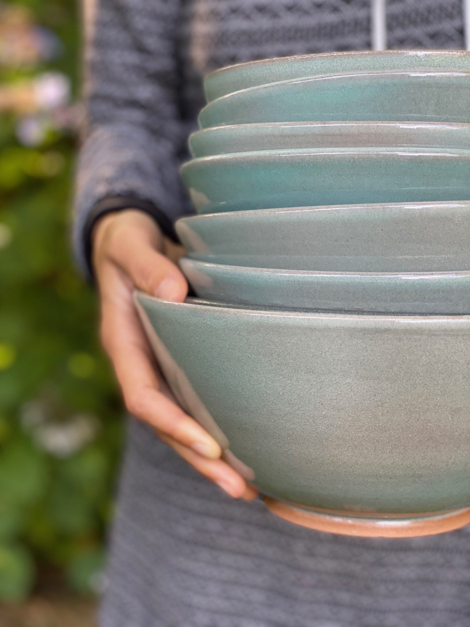 Person holding a stack of teal ceramic bowls outdoors, blurred background with greenery. Sonia Lesage Ceramics