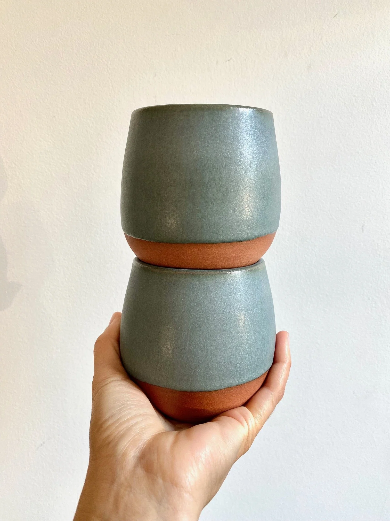 A person holding a stack of matte ceramic cups, each with a rounded shape and a combination of muted blue/green and terracotta colors, against a plain off-white wall background. Sonia Lesage Ceramics