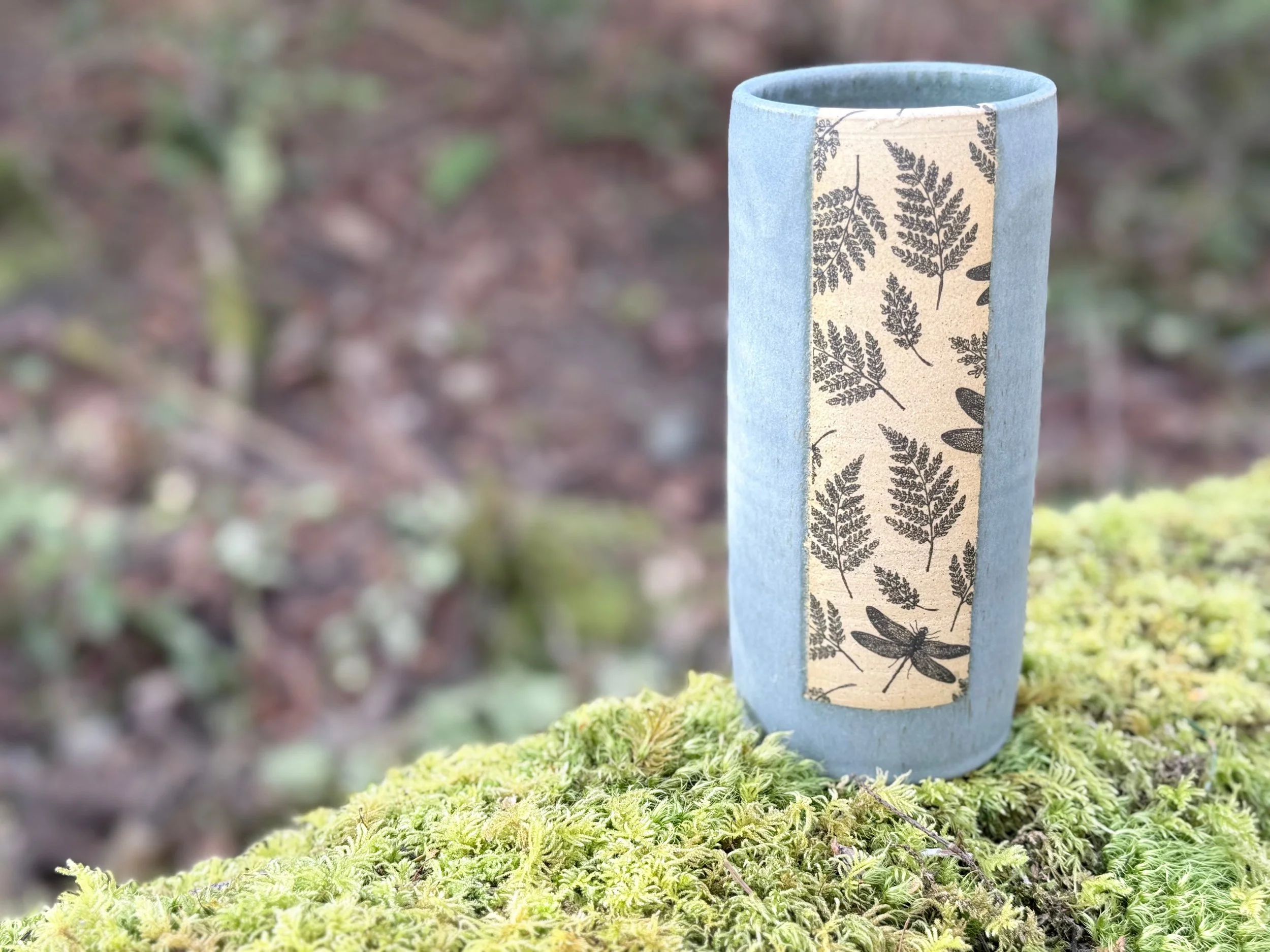A tall, cylindrical, blue ceramic vase with an underglaze transfer featuring black fern and dragonfly illustrations, placed on moss-covered ground outdoors. Sonia Lesage Ceramics