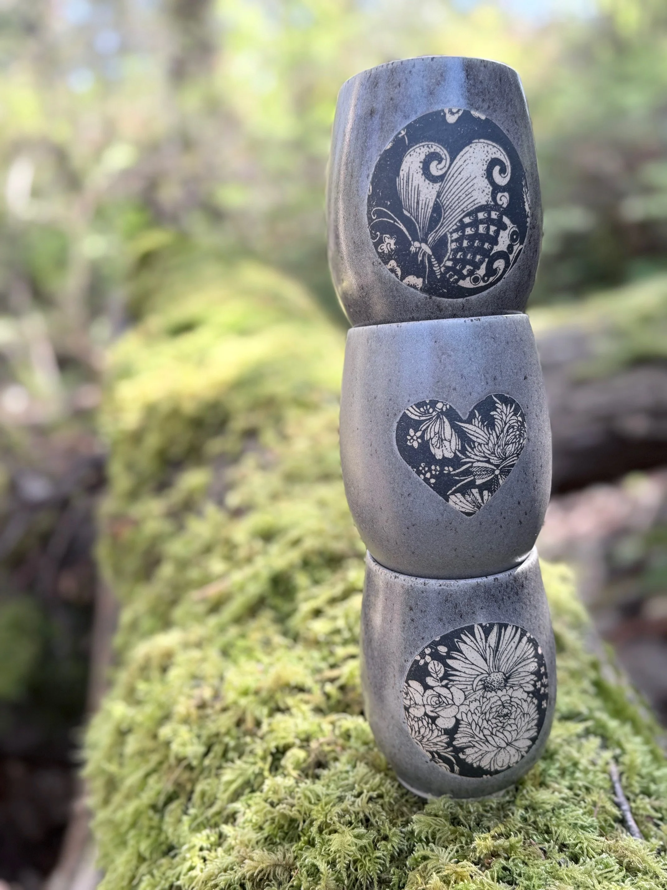 Stacked grey/brown tumblers with black floral and heart patterns on their surfaces, outdoors on mossy ground. Sonia Lesage Ceramics