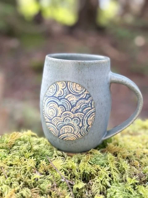 A ceramic mug with a swirl pattern on its front, placed outdoors on moss with trees in the background. Stormy skies/waves mug - Sonia Lesage Ceramics