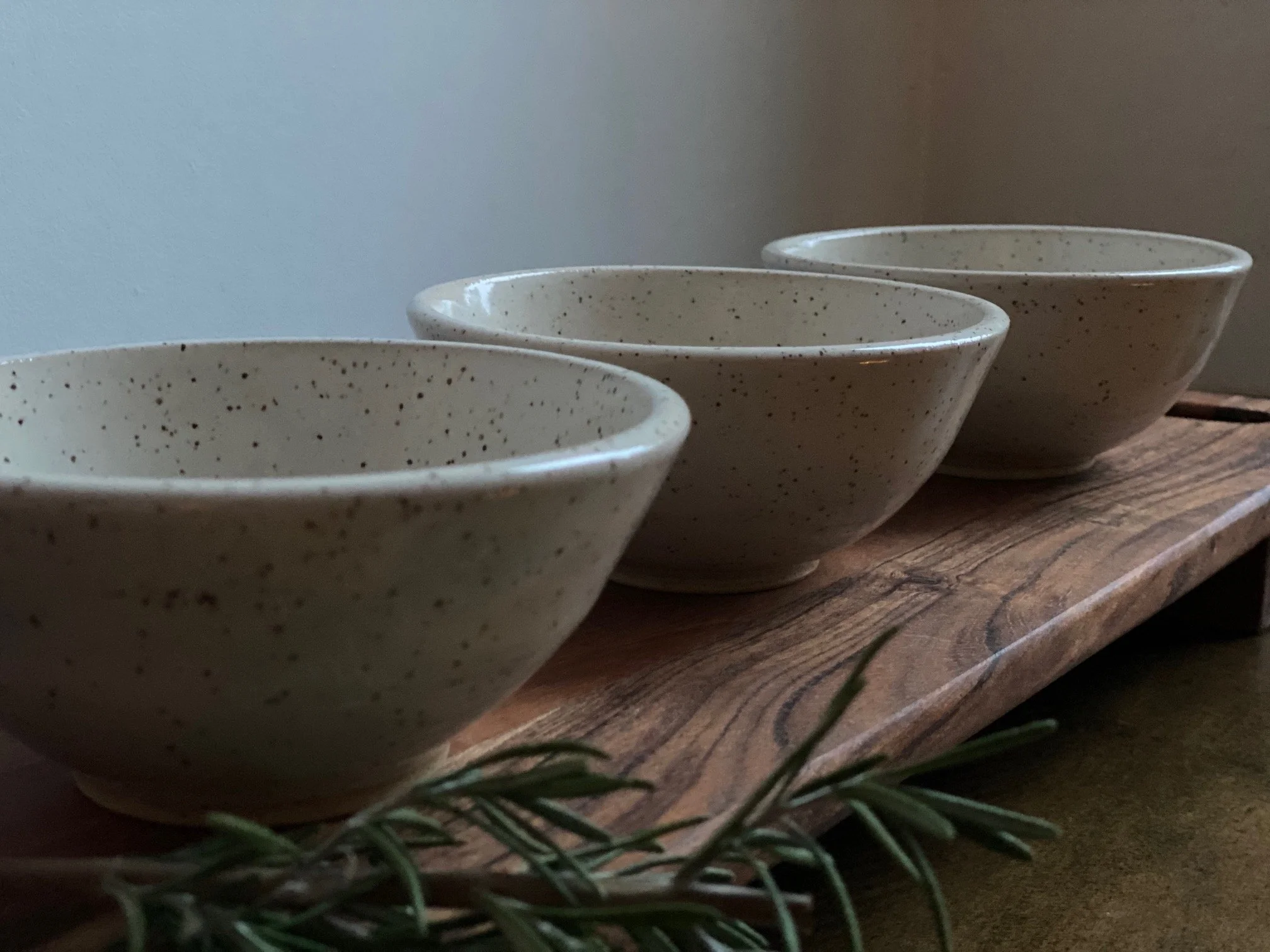 Three speckled beige ceramic bowls on a wooden board with a sprig of green foliage in the foreground. Sonia Lesage Ceramics