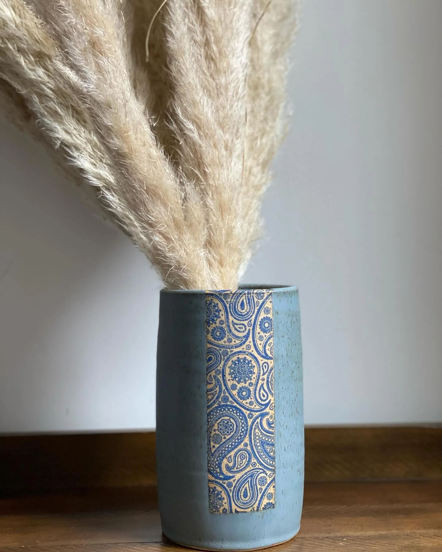 Beige dried pampas grass in a blue ceramic vase with a decorative blue pattern, placed on a wooden surface against a plain wall. Sonia Lesage Ceramics