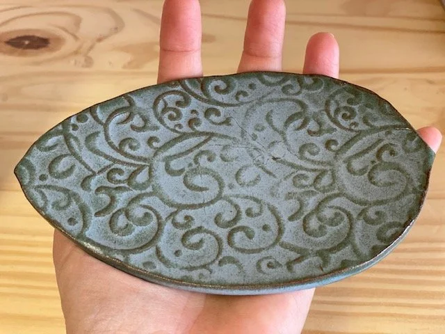 Hand holding a decorative ceramic leaf plate with greenish-blue glaze and a swirling vine pattern, oval shape, against a wooden background. Sonia Lesage Ceramics