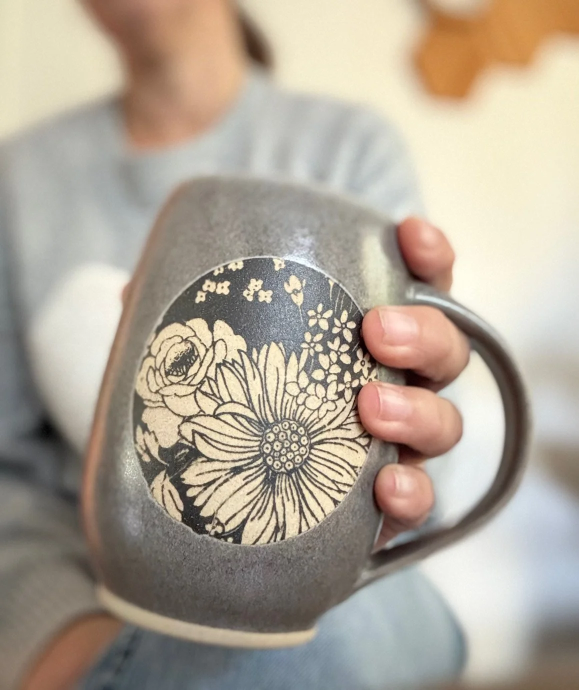 Close-up of a brown/gray mug with an underglazed floral design, held by a person in a light-colored sweater. Sonia Lesage Ceramics