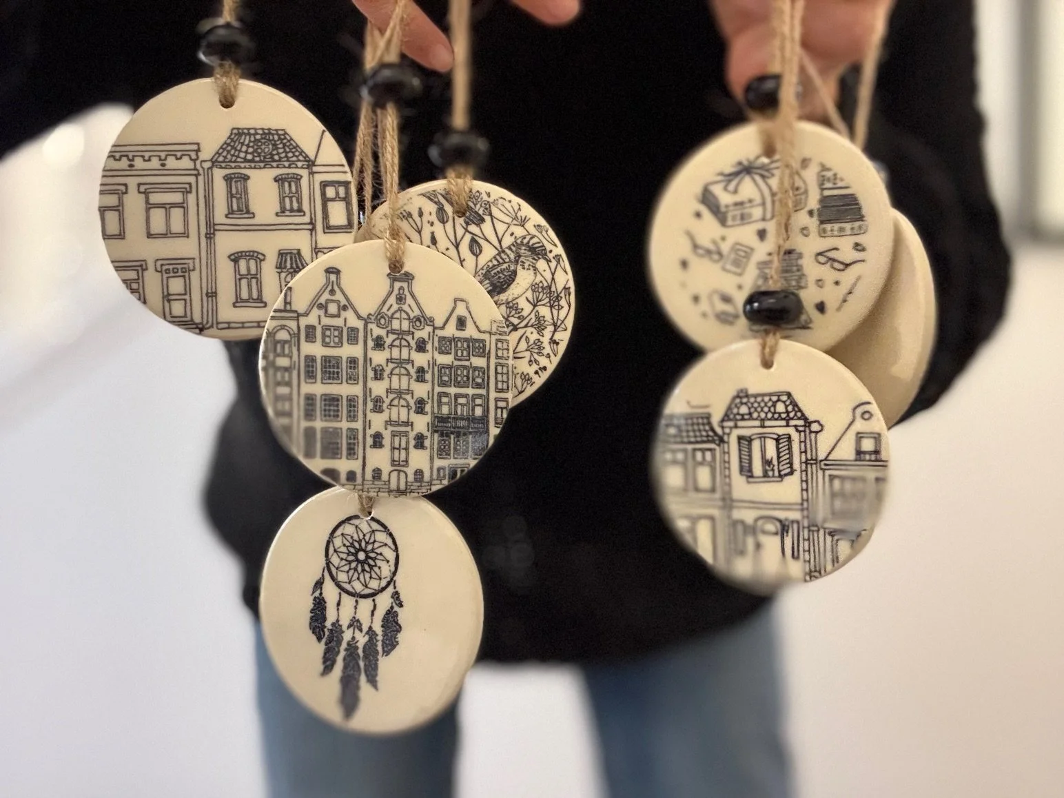 A person holding a round, cream-colored ceramic ornaments with various designs including European towns, a bird, and a dreamcatcher. The background is blurred. Sonia Lesage Ceramics