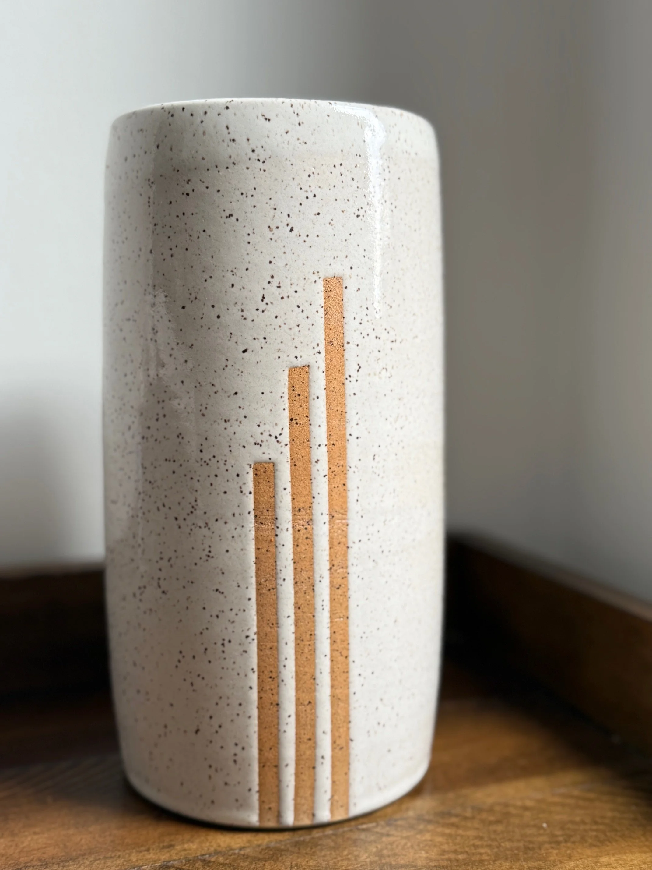 A ceramic vase with a speckled off-white surface and three vertical stripes  of exposed clay on it, placed on a wooden surface. Sonia Lesage Ceramics