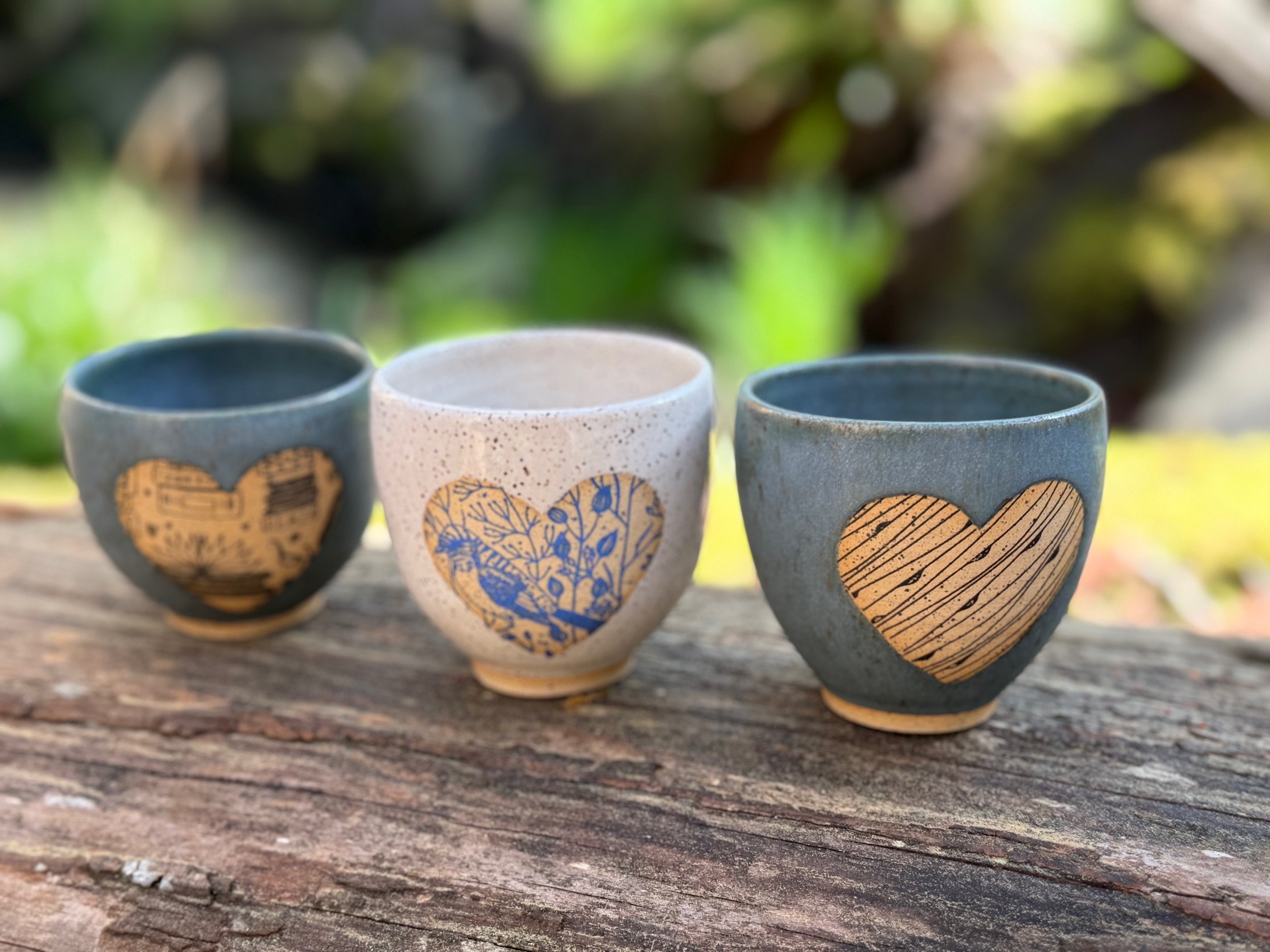 Three small espressi/tea cups with heart-shaped designs on a weathered wooden surface outdoors, with a blurred green and natural background. Sonia Lesage Ceramics