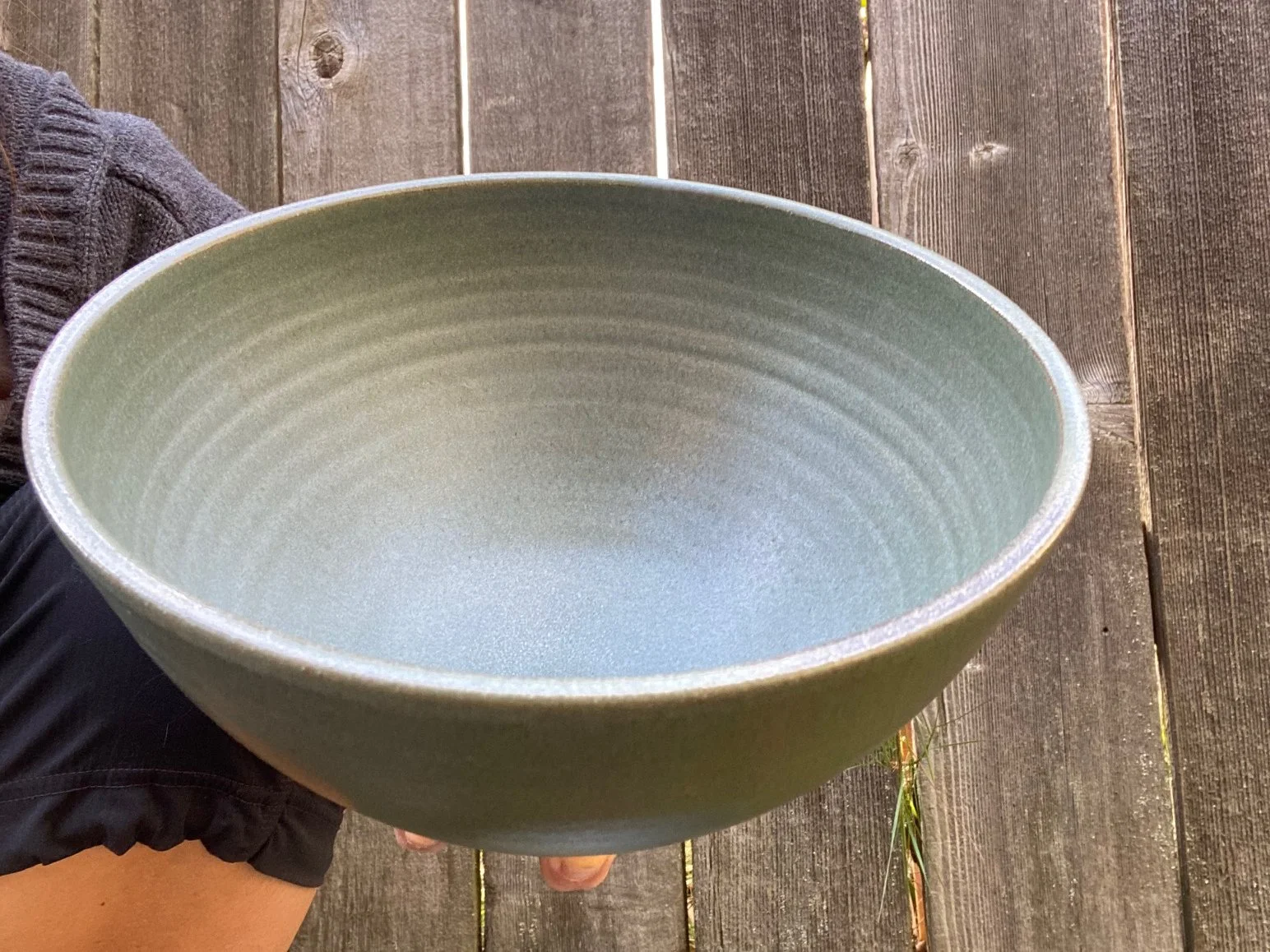 Person holding a large blue/grey ceramic bowl with concentric rings, sitting outdoors on a wooden surface. Sonia Lesage Ceramics
