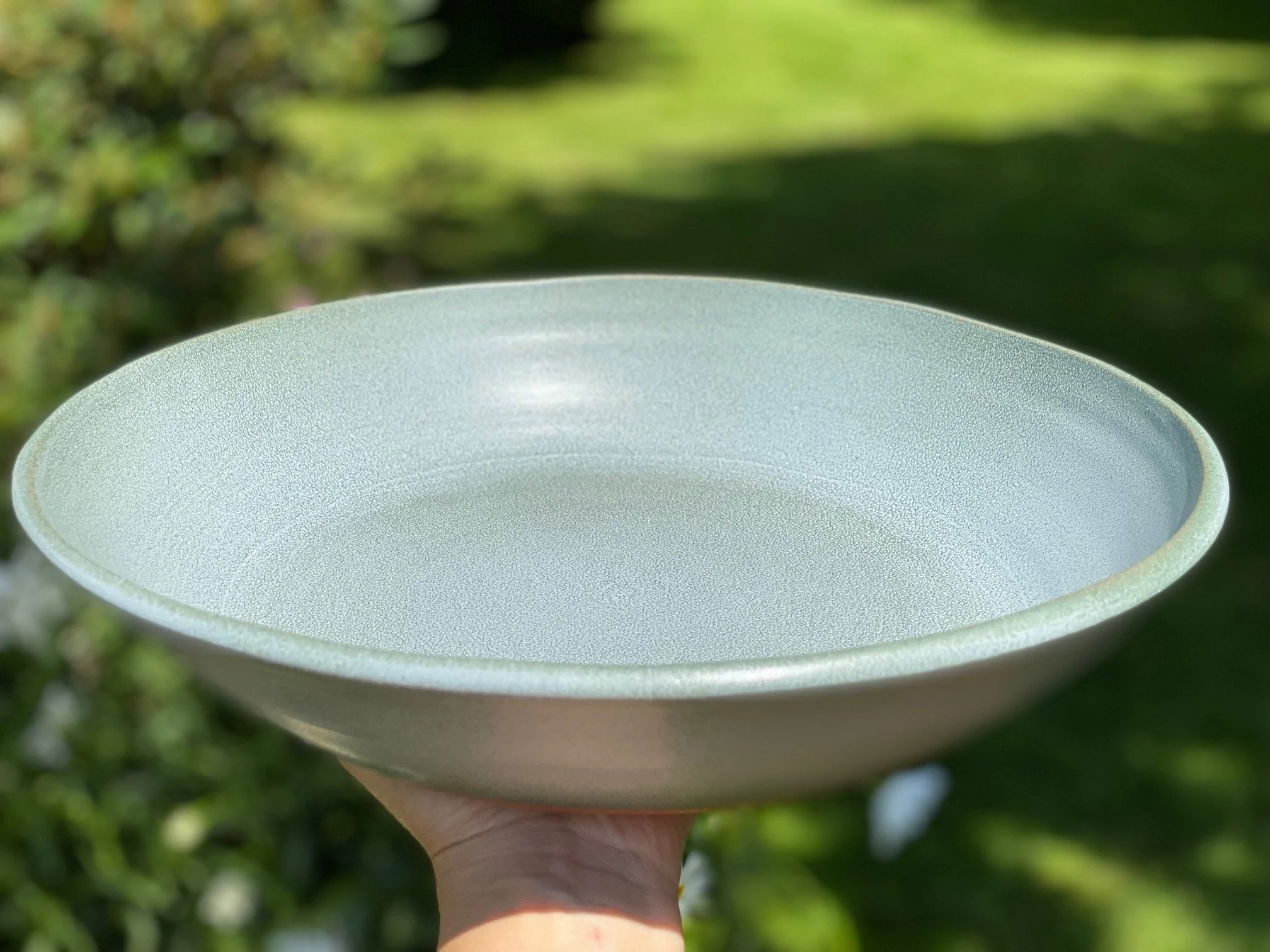 A person holding a large blue/gray platter outdoors with greenery in the background. Sonia Lesage Ceramics