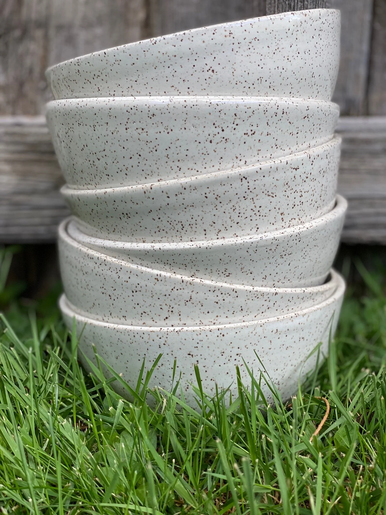 A stack of five speckled white ceramic bowls, placed on grass outdoors with a weathered wooden fence in the background. Sonia Lesage Ceramics