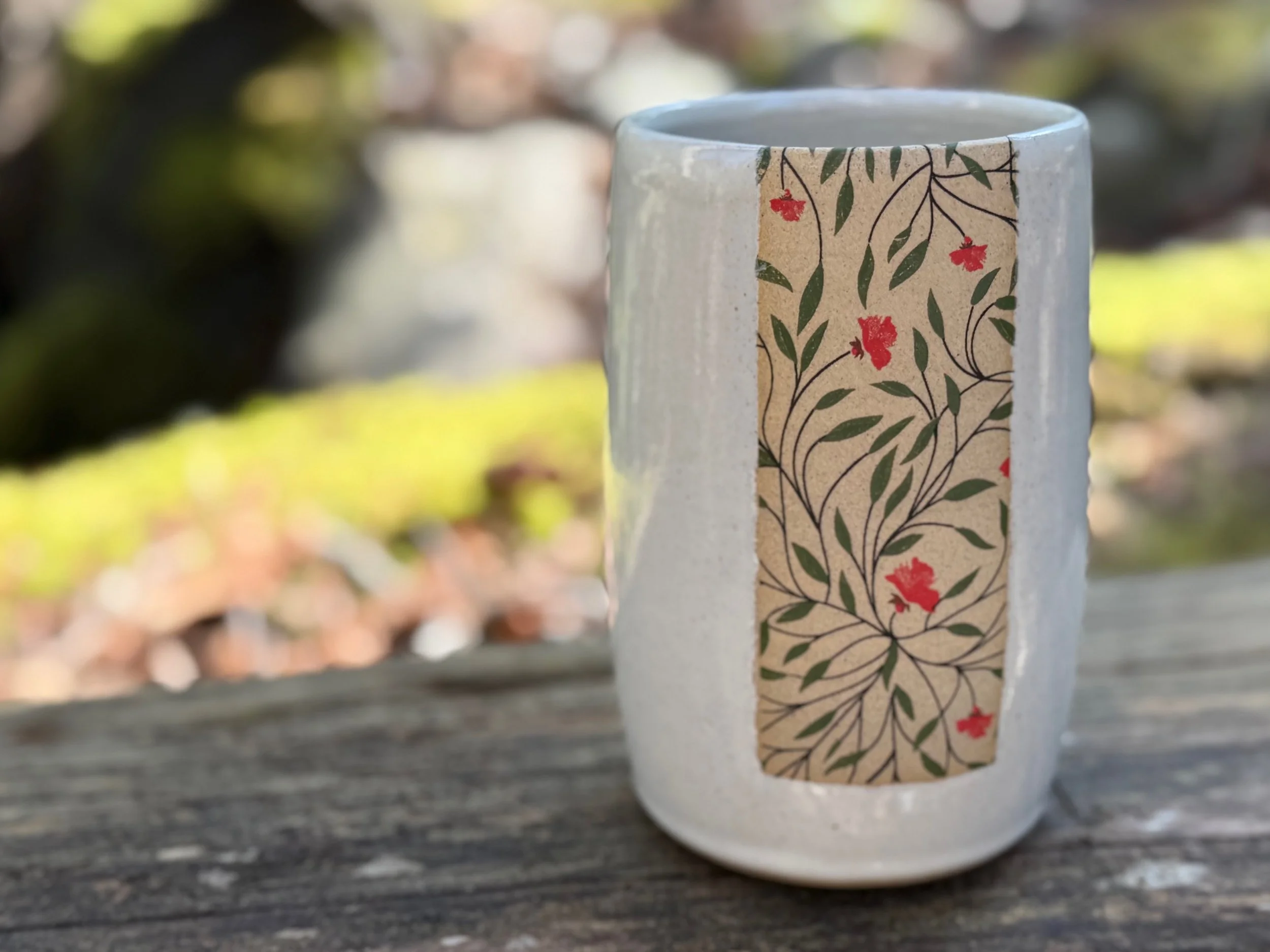 A ceramic vase with a decorative floral pattern, placed on a wooden surface outdoors, with a blurry natural background. Sonia Lesage Ceramics