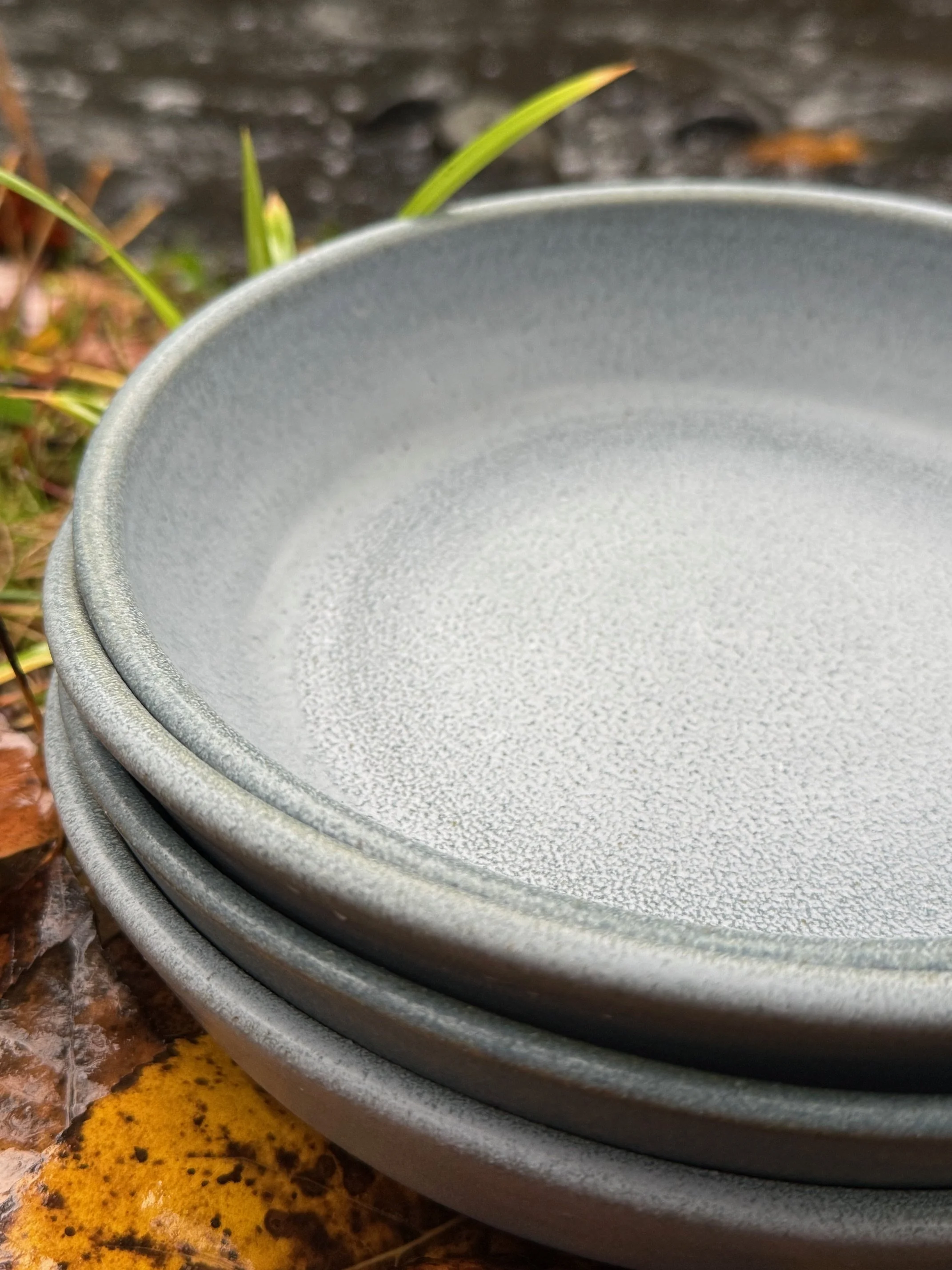 Stacked blue/gray ceramic platters outdoors on a brown fallen leaves ground with green grass and a dark wooden surface in the background. Sonia Lesage Ceramics