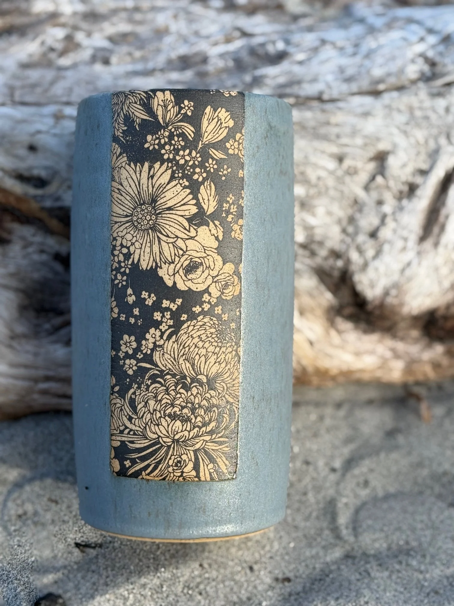 A vase with a floral patterned strip, set against a beach background with sand and driftwood. Sonia Lesage Ceramics