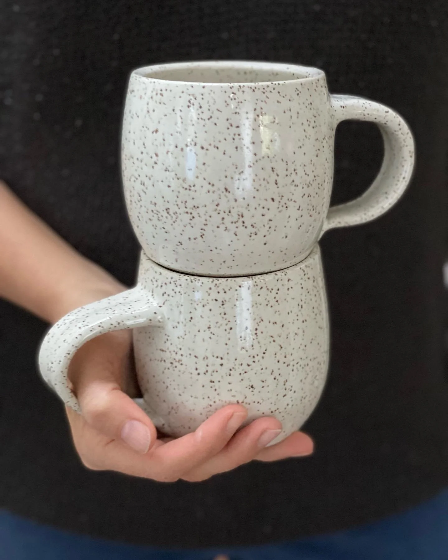Two speckled ceramic mugs stacked, one on top of the other, with a person holding them against a dark background. Sonia Lesage Ceramics