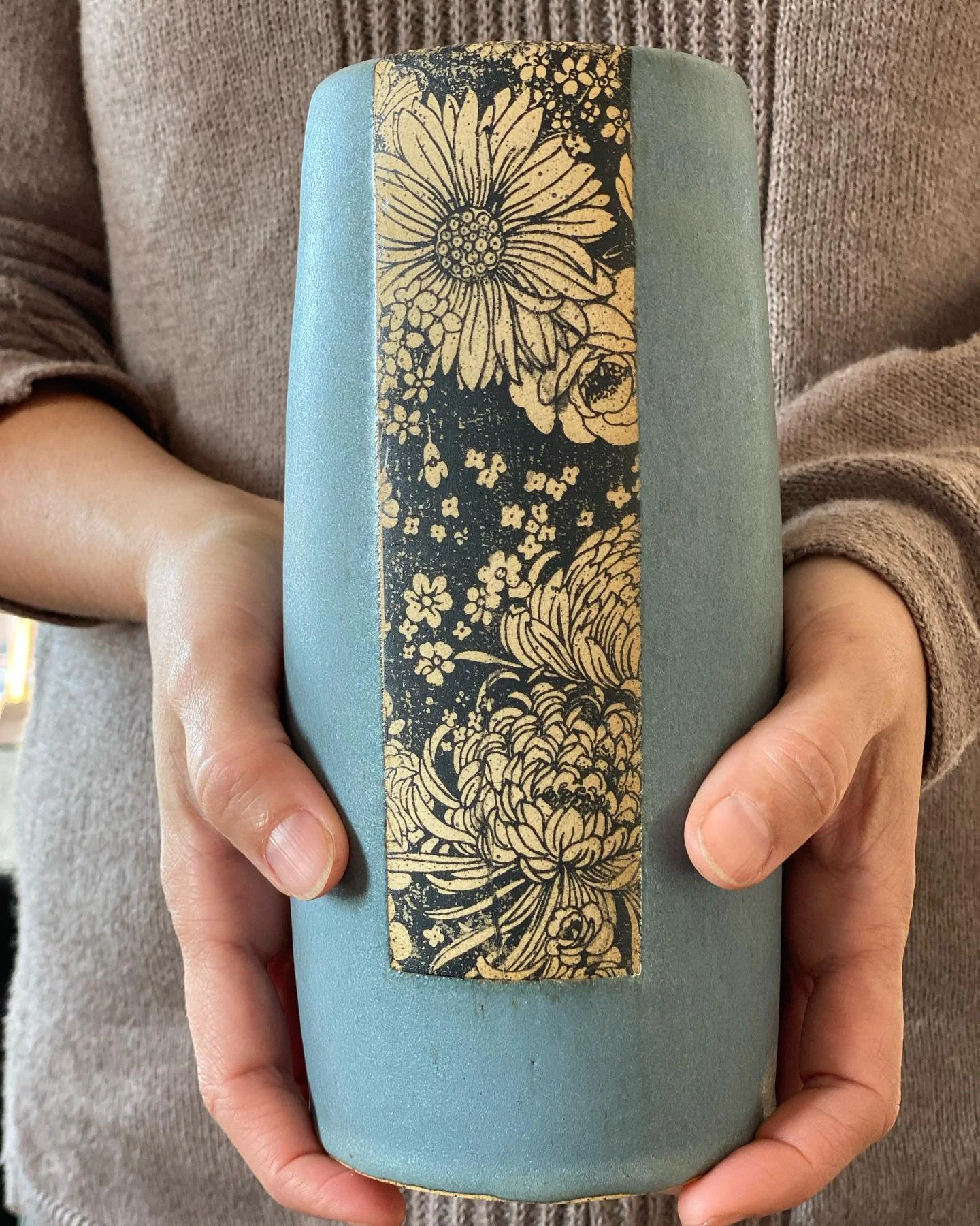 Person holding a vase with a floral design in beige and black, featuring large flowers and smaller blossoms. Sonia Lesage Ceramics