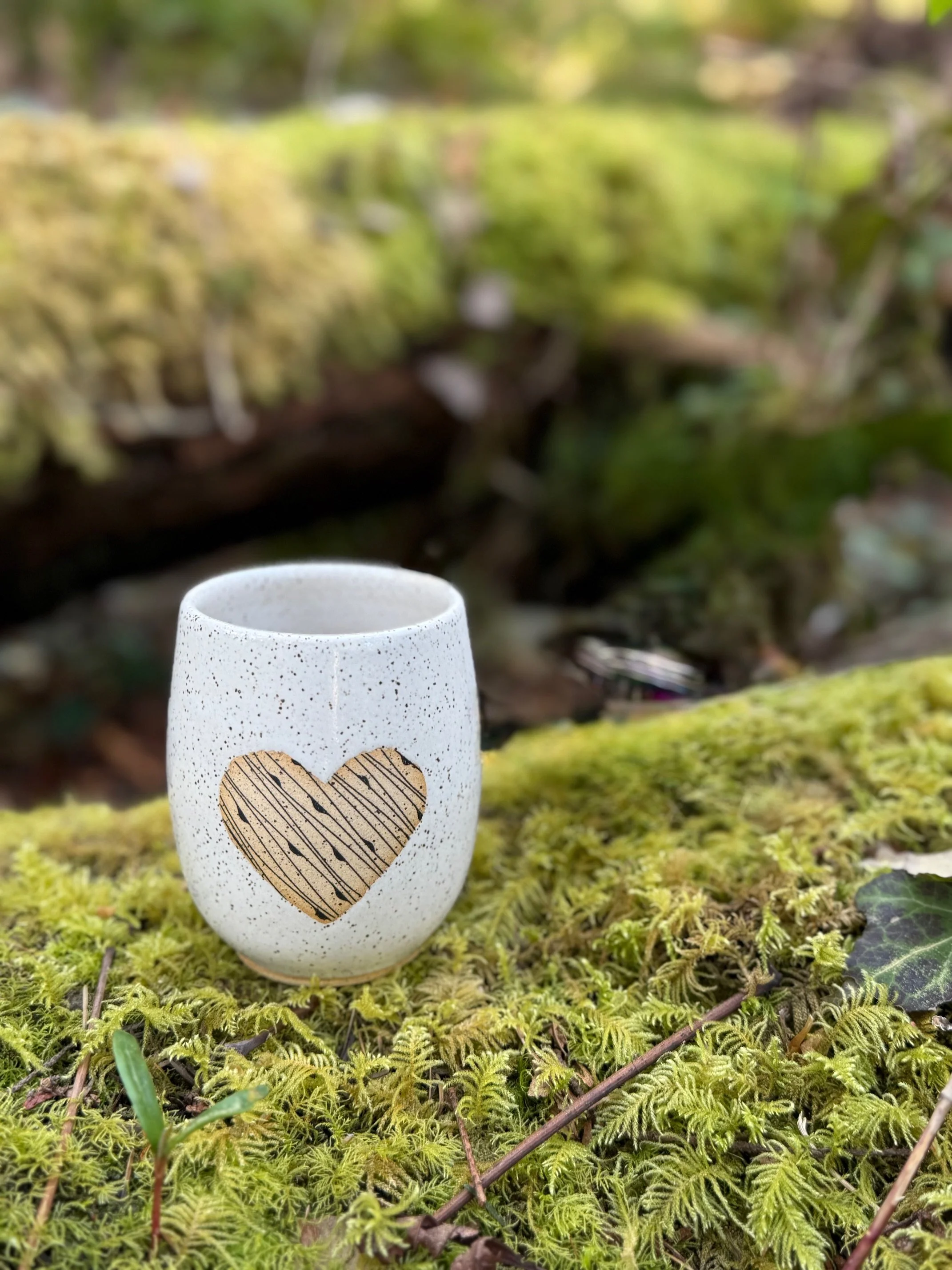 Speckled white tumbler with a heart design, placed on moss-covered ground in a forest setting. - Sonia Lesage Ceramics