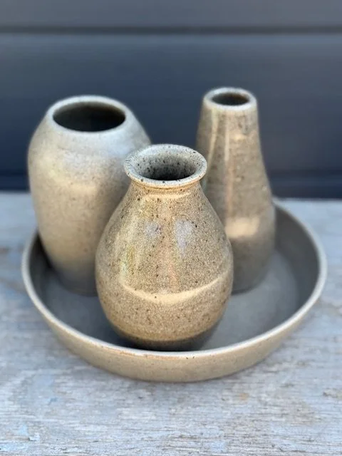 Three small beige ceramic bud vases on a round tray, placed on a wooden surface with a dark background. Sonia Lesage Ceramics