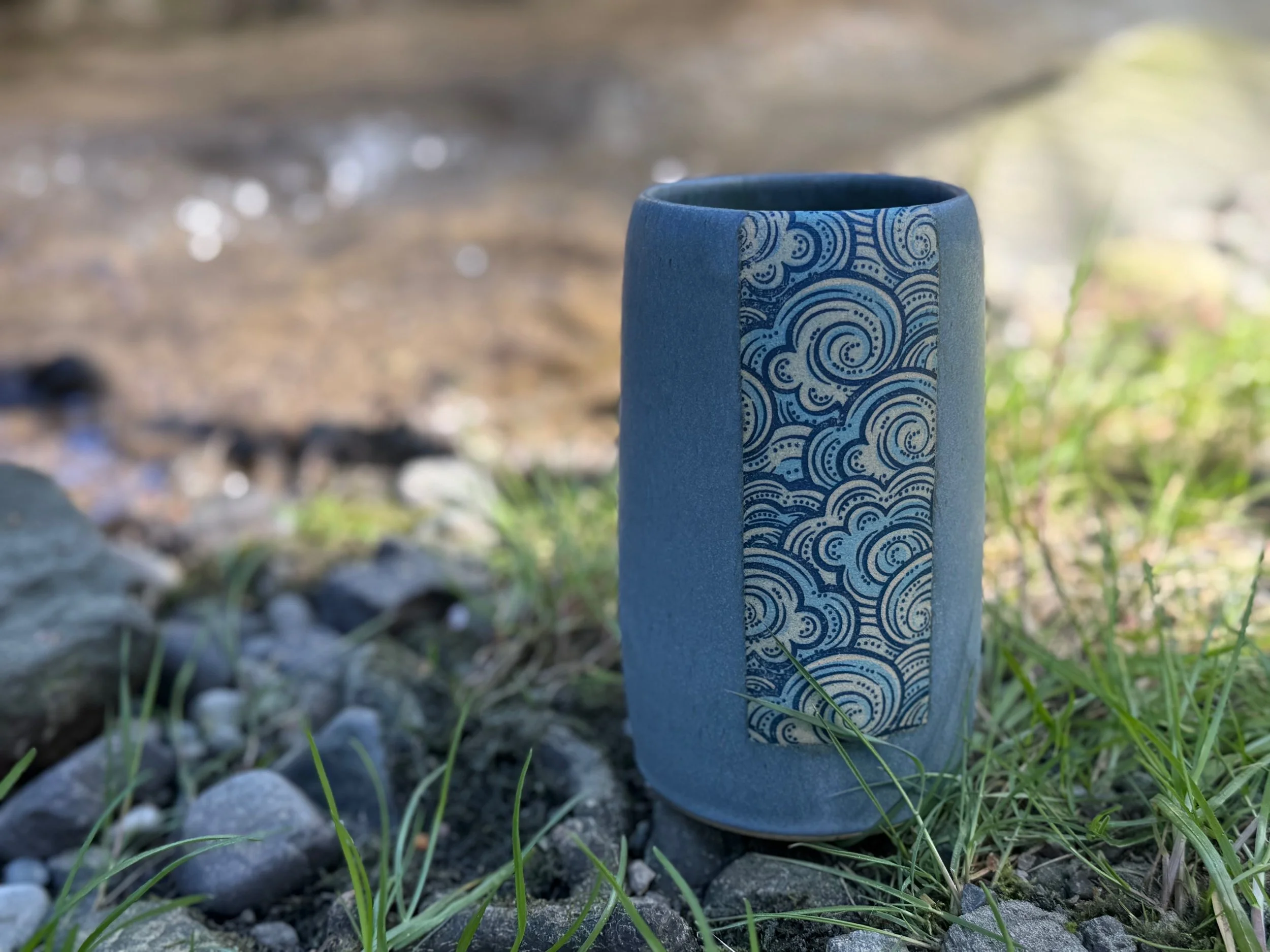 A blue ceramic vase with a decorative panel featuring blue and white swirling patterns, placed outdoors on rocks and grass with a blurred natural background. Sonia Lesage Ceramics