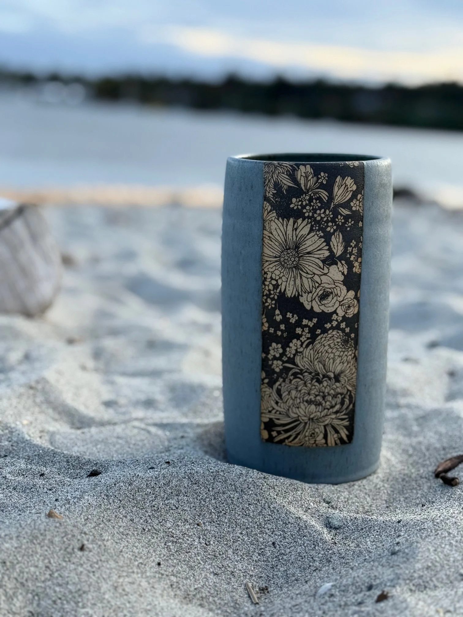A ceramic vase with a floral pattern on a sandy beach, with water and distant land in the background. Sonia Lesage Ceramics
