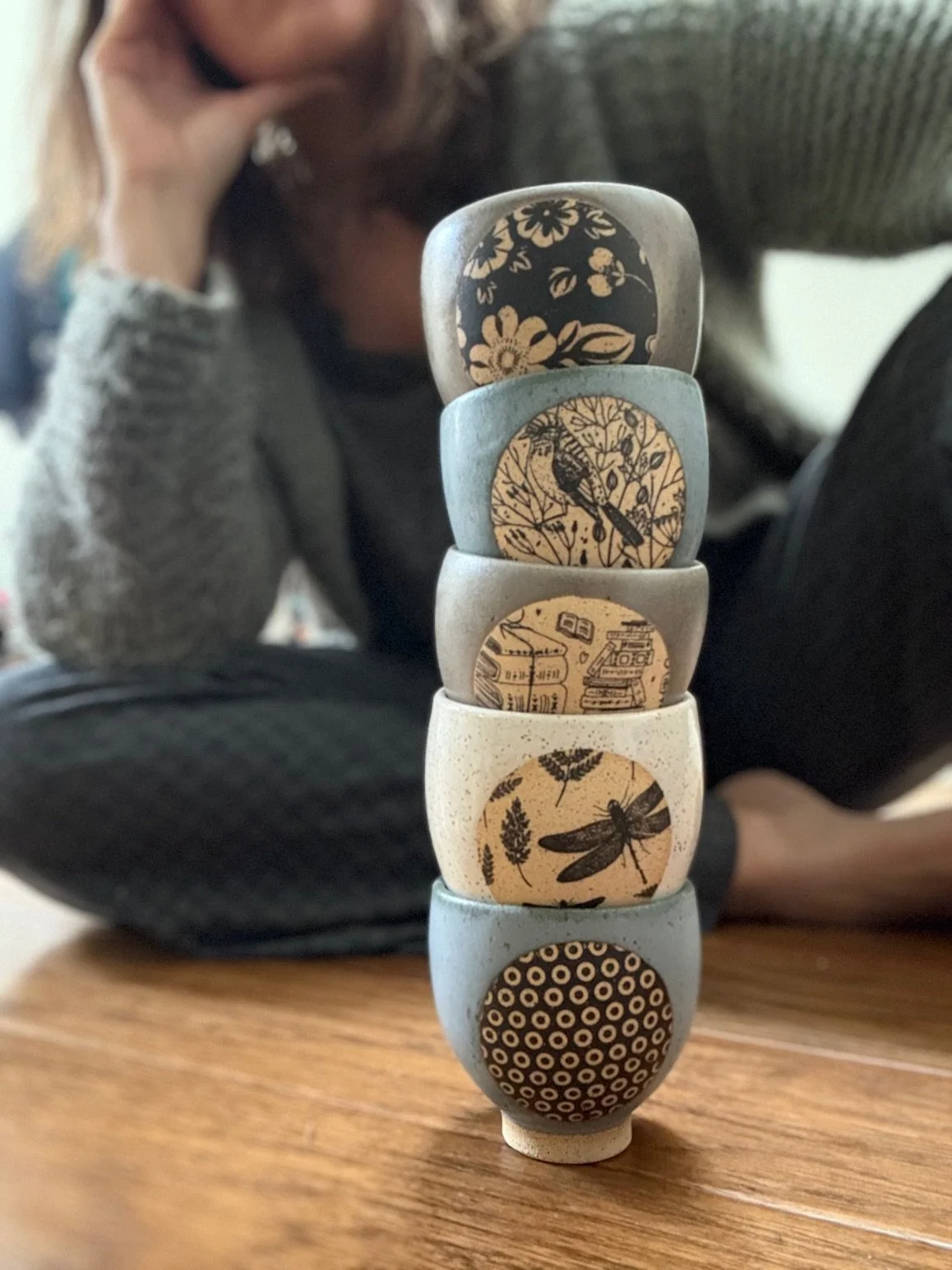 Stacked espresso/tea cups with underglaze transfer designs, placed on a wooden surface, with a person sitting in the background. Sonia Lesage Ceramics