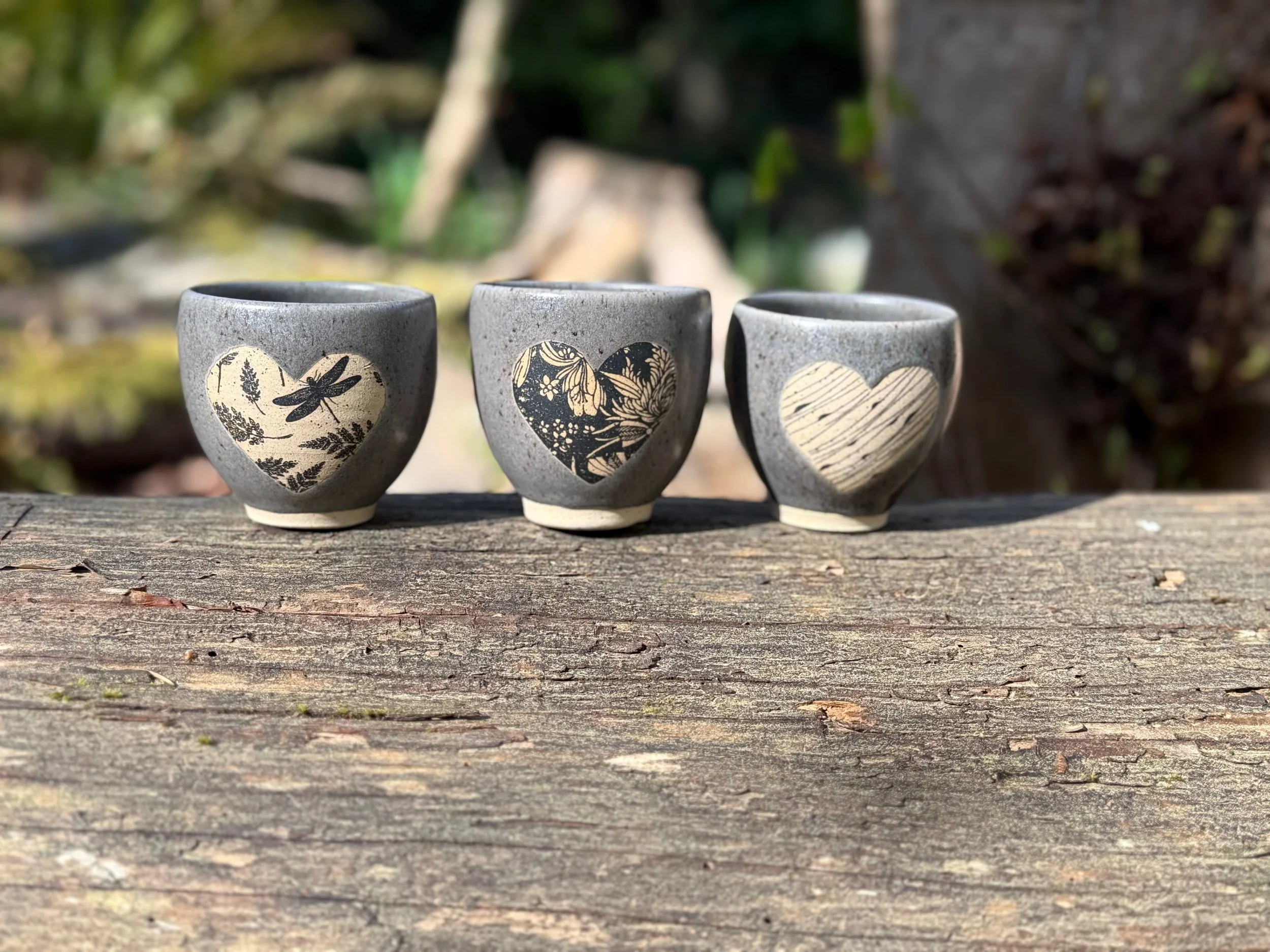 Three small ceramic espresso/tea cups with underglaze transfer designs, placed on a wooden surface outdoors with blurred trees and plants in the background. Sonia Lesage Ceramics