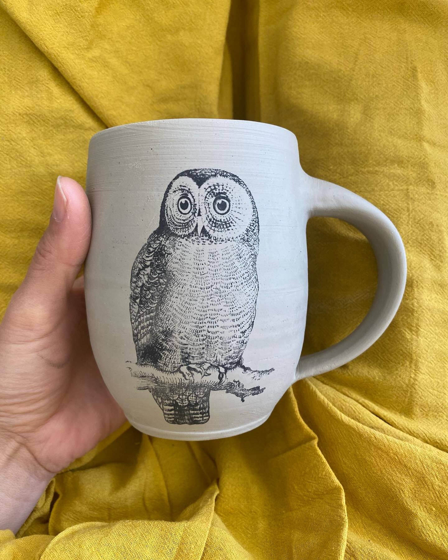 A ceramic mug with a black illustration of an owl on its side, held by a person's hand, against a yellow fabric background. Sonia Lesage Ceramics