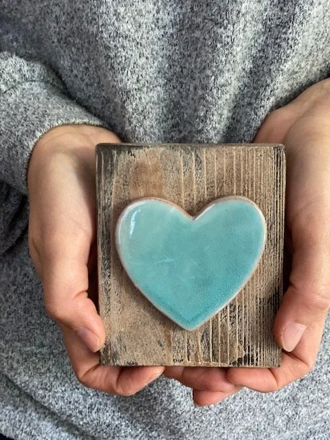Person holding a wooden block with a heart-shaped blue ceramic piece on top. Sonia Lesage Ceramics