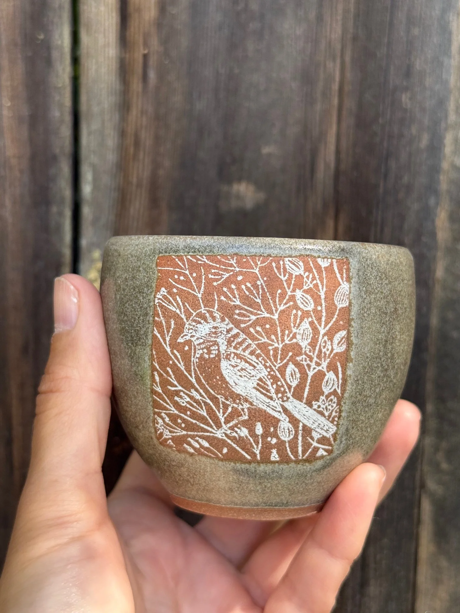 Ceramic cup with a white outlining image of a bird and foliage on a brown background, held in a person's hand against a wooden background. Sonia Lesage Ceramics