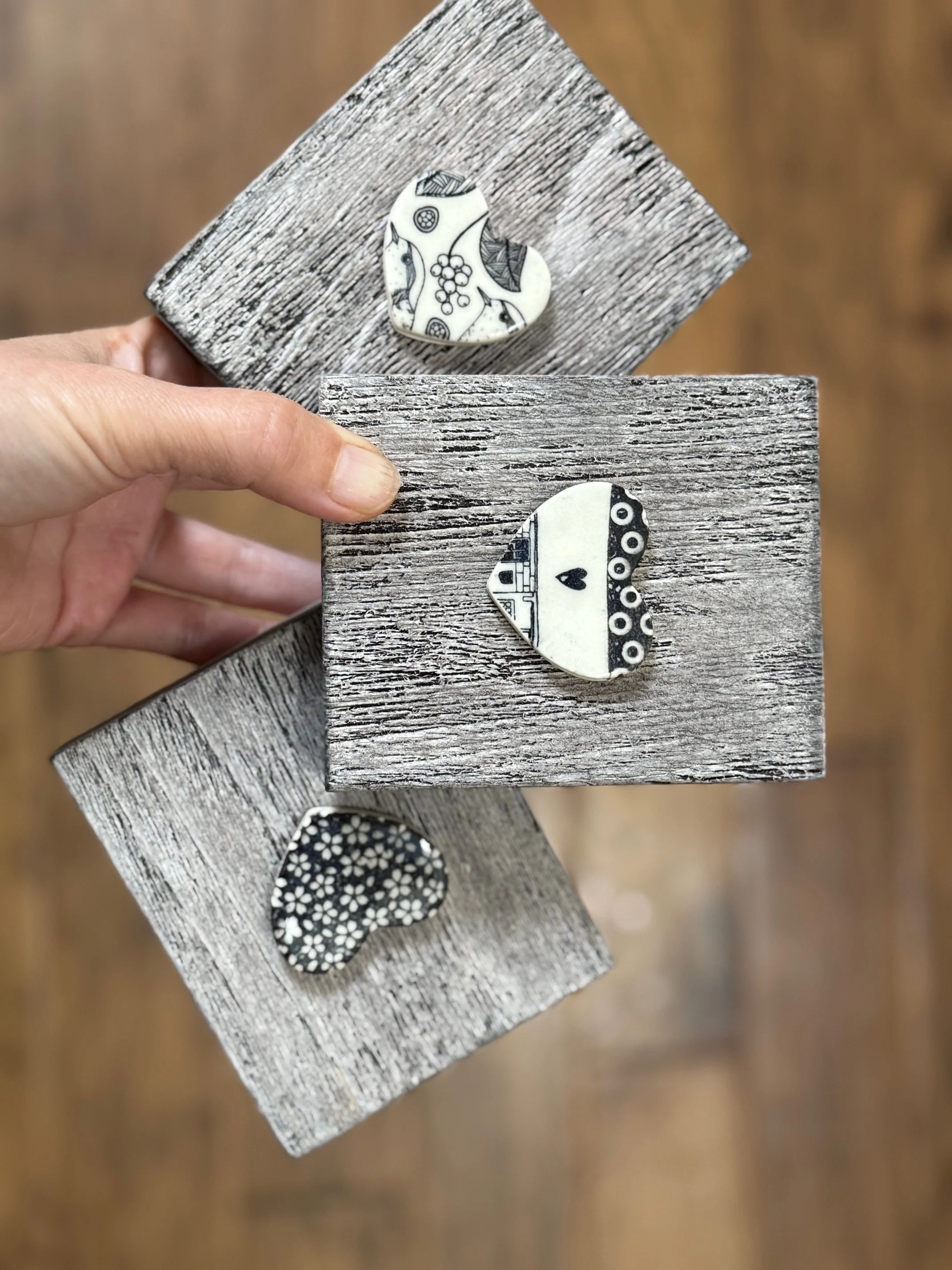 Three wall hangings with decorative heart-shaped ceramic pieces, some with floral or patterned designs, held by a hand against a blurred wood floor background. Sonia Lesage Ceramics