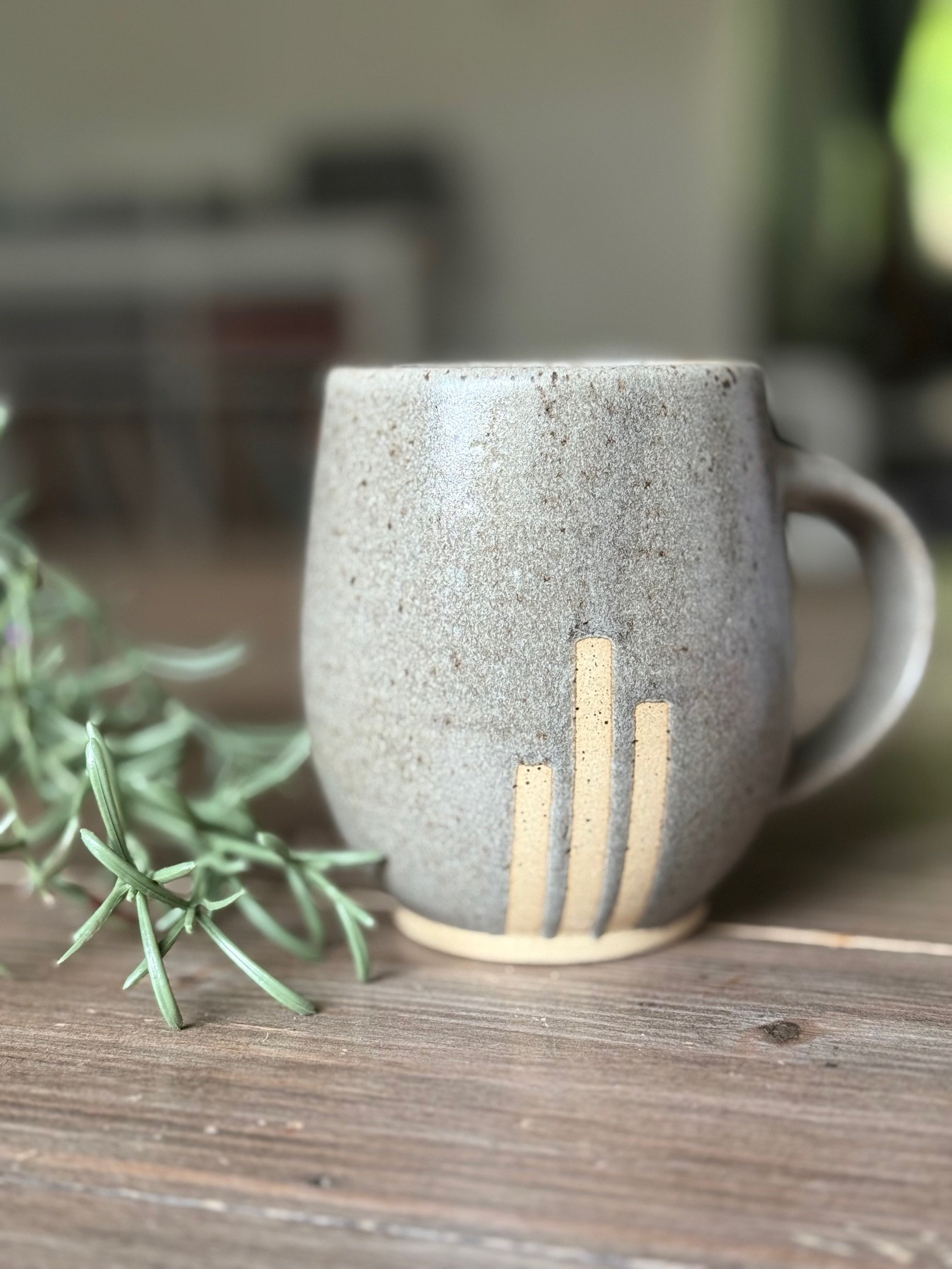 A ceramic mug with a textured brown/gray surface and vertical stripes of exposed clay, on a wooden table, with a sprig of green herb beside it, and a blurred background. Sonia Lesage Ceramics