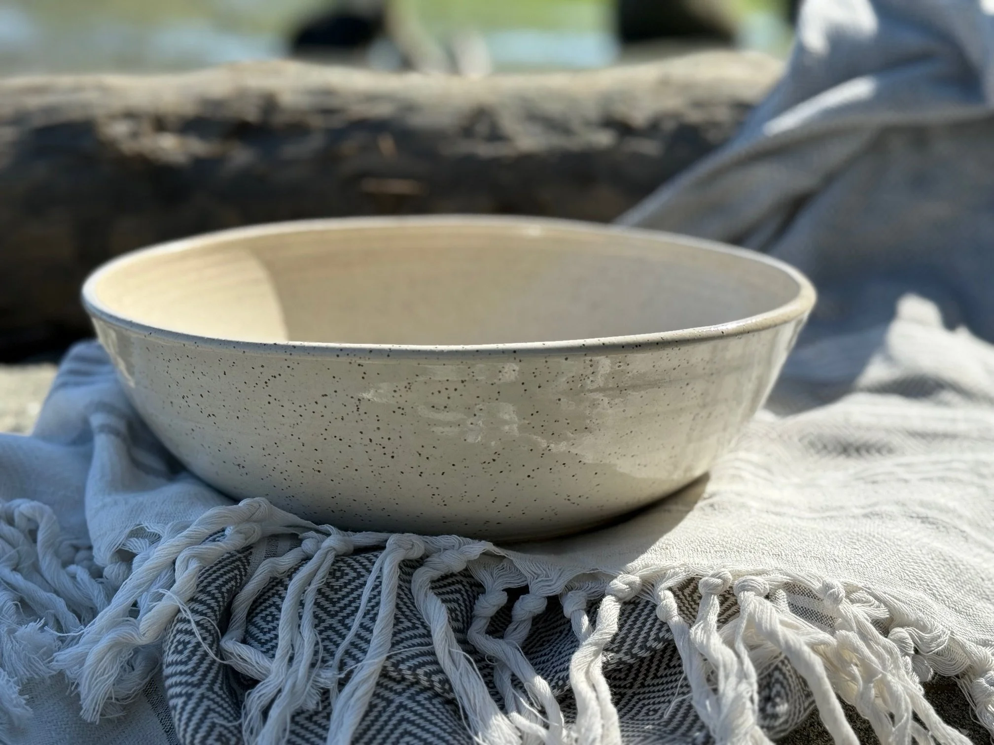 A ceramic bowl with white speckled glaze, resting on a fringed cloth outdoors. Sonia Lesage Ceramics