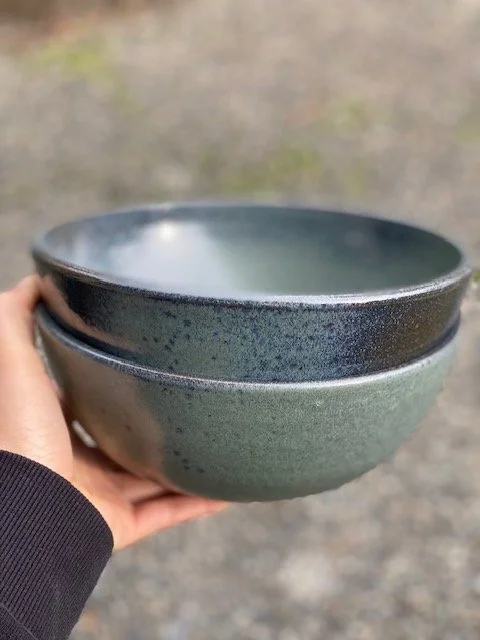Stacked blue ceramic bowls held in a person's hand outdoors, with a blurry natural background. Sonia Lesage Ceramics