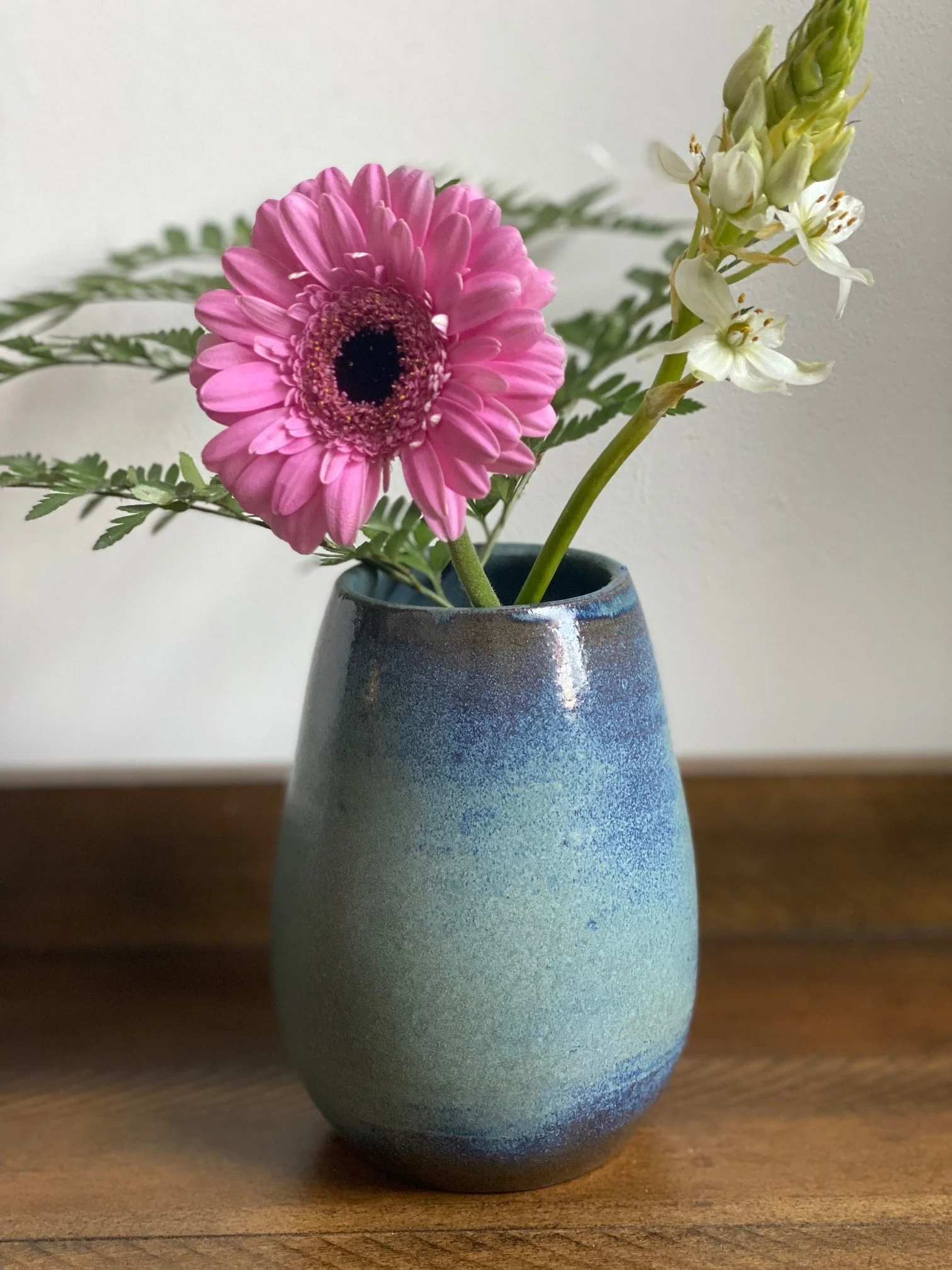 A ceramic vase with a blue-green gradient, containing a pink gerbera daisy, white flowers, and green foliage, placed on a wooden surface. Sonia Lesage Ceramics