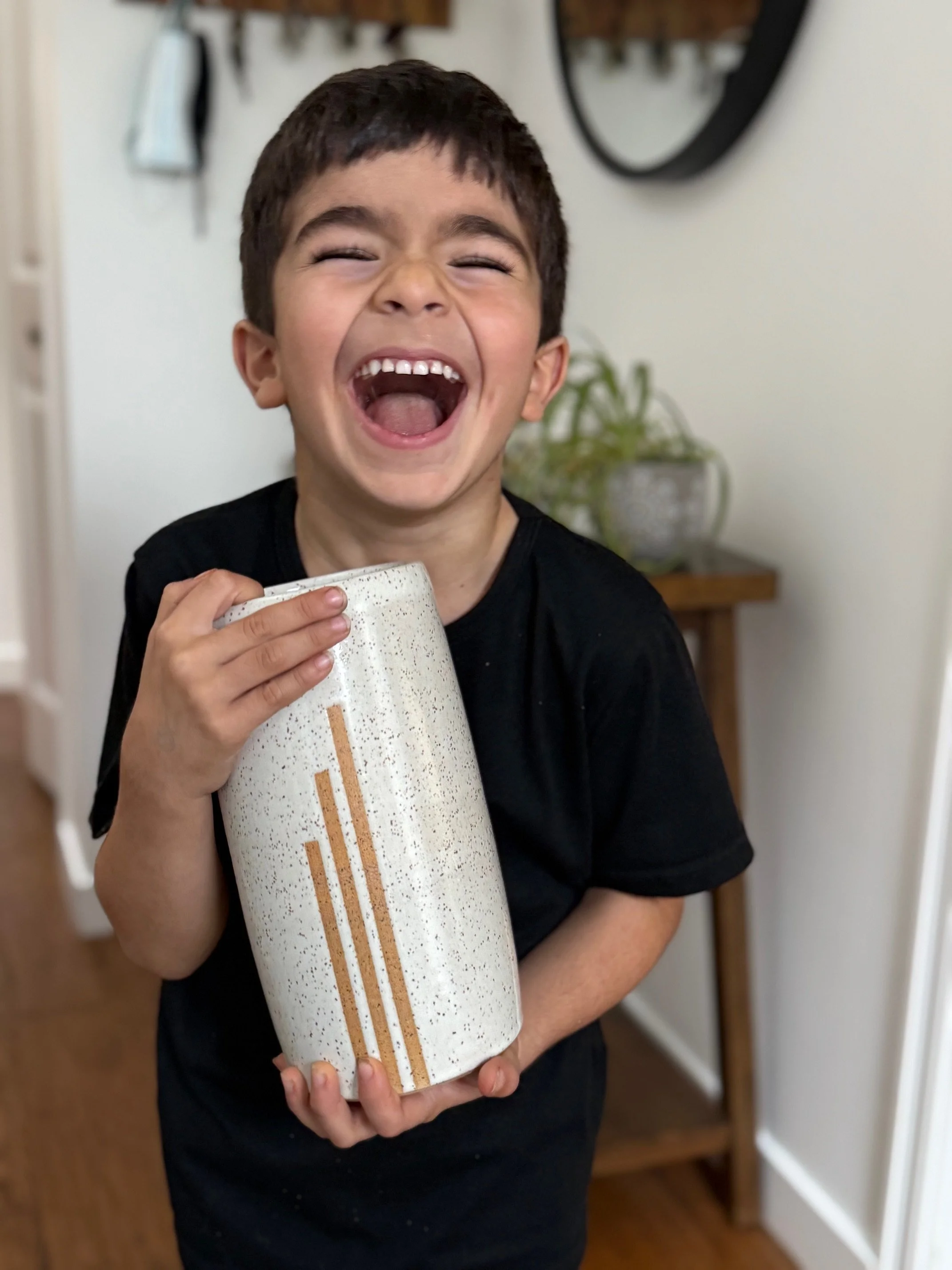 A young boy with short dark hair, wearing a black shirt, is laughing joyfully and holding a white ceramic vase with a speckled pattern and three vertical brown stripes. The background shows a small table with a potted plant and a wall with some hangi