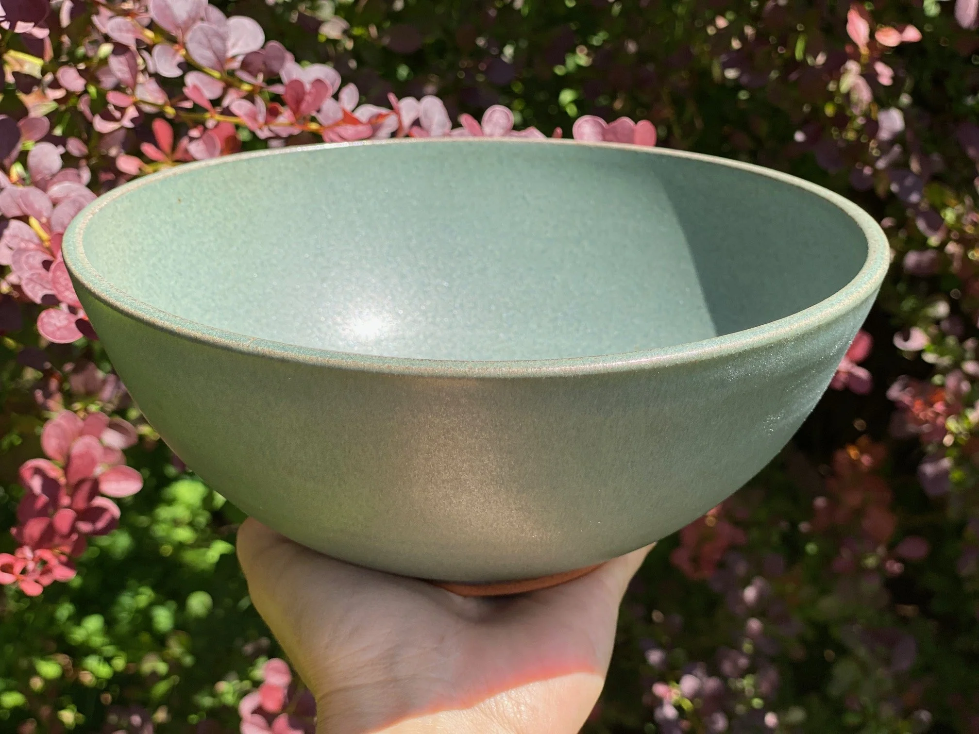 A hand holding a large, blue/grey ceramic bowl outdoors, with pink and purple flowers in the background. Sonia Lesage Ceramics