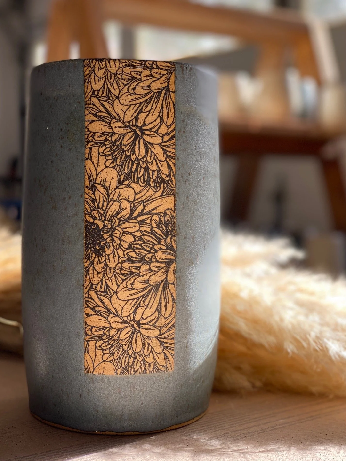 Close-up of a blue/grey ceramic vase with a vertical strip with an underglazed transfer of a bird illustration, placed on a wooden surface with a fluffy pampas grass nearby. Blurred wooden furniture in the background. Sonia Lesage Ceramics