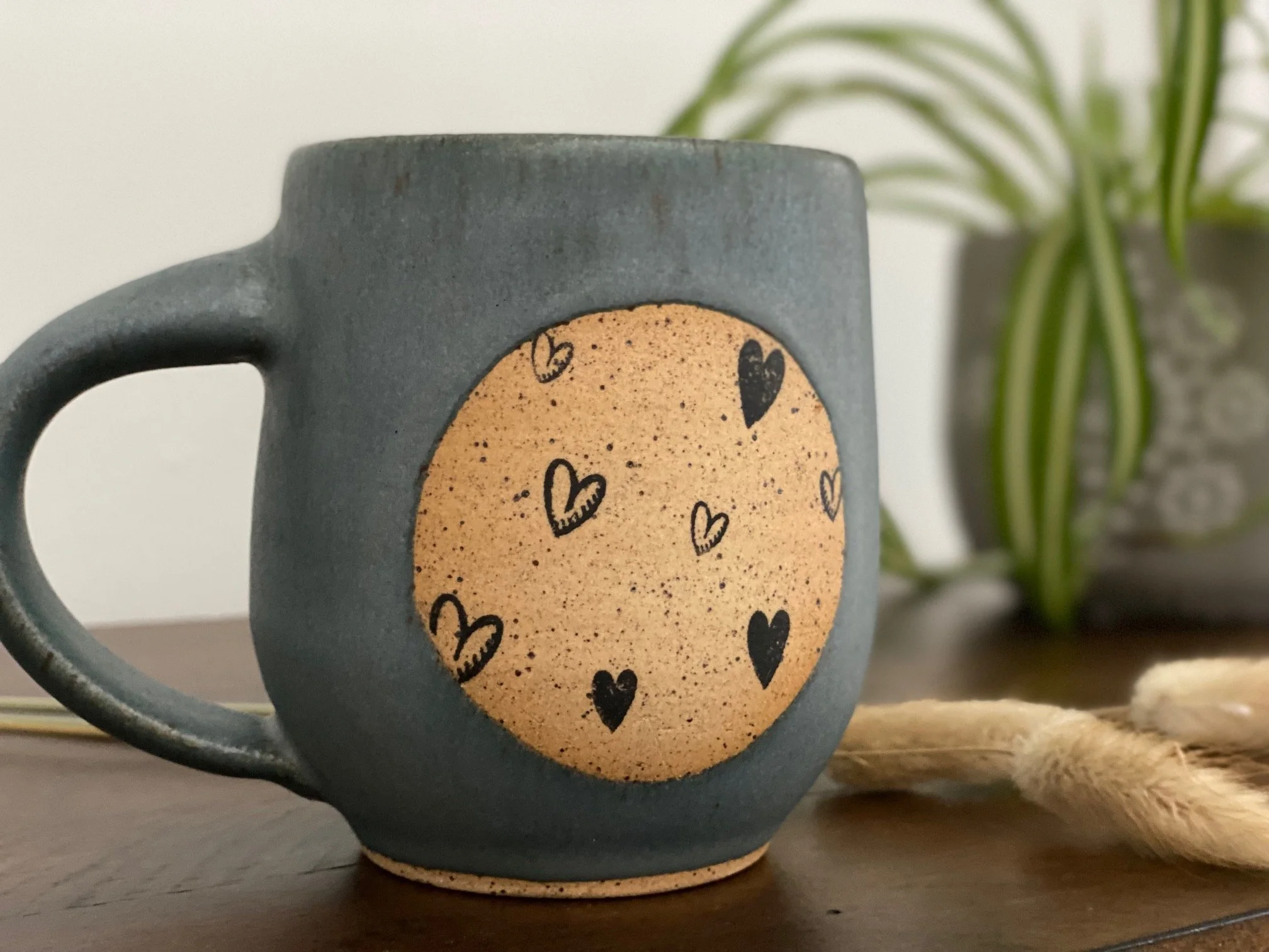 A ceramic mug with a blue-gray exterior and a beige circular design decorated with black hearts on its surface. In the background, there is a blurred potted plant with long, green leaves. Sonia Lesage Ceramics