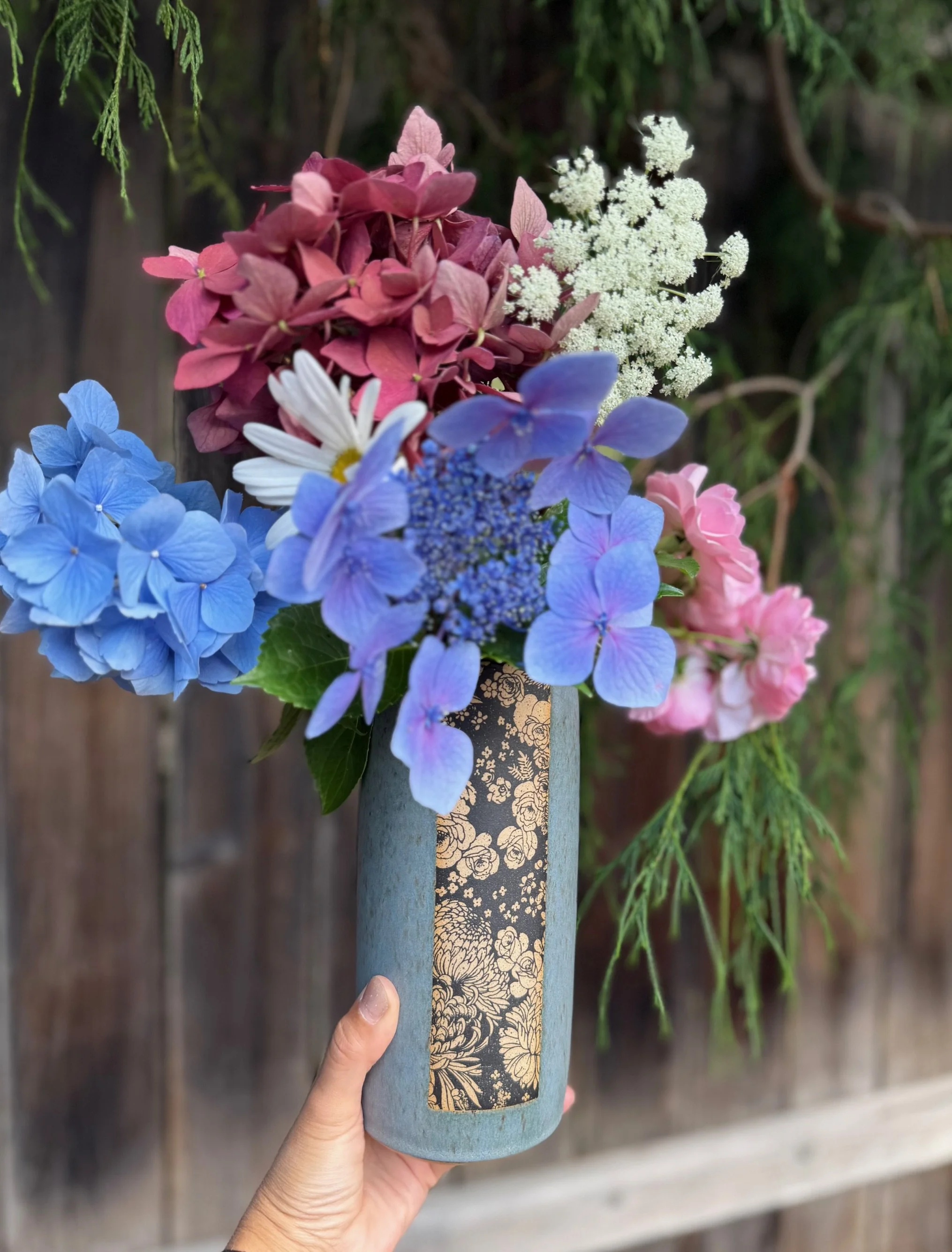 A hand holds a ceramic vase with a floral design, filled with a colorful bouquet of pink, blue, white, and purple flowers, against a wooden fence and green foliage background. Sonia Lesage Ceramics