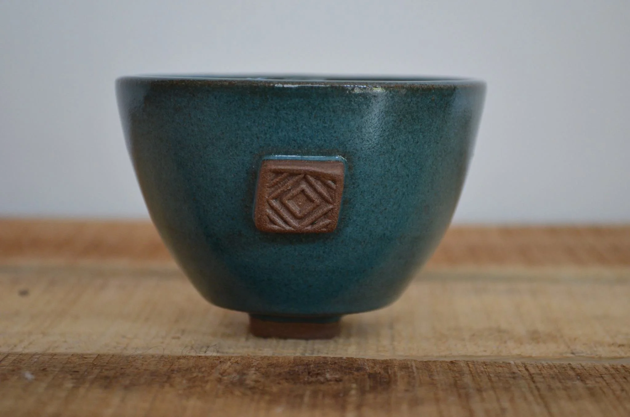 A ceramic cup with a textured dark teal glaze and a small brown square emblem on the side, placed on a wooden surface. Sonia Lesage Ceramics