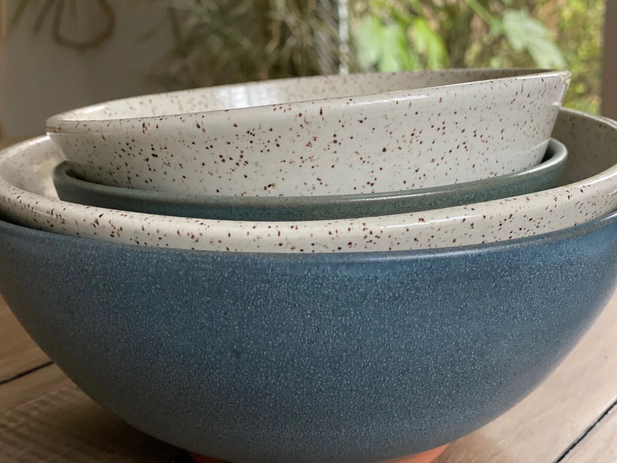 Stack of ceramic bowls in various shades, including white with speckles, and blue, on a wooden surface. Sonia Lesage Ceramics