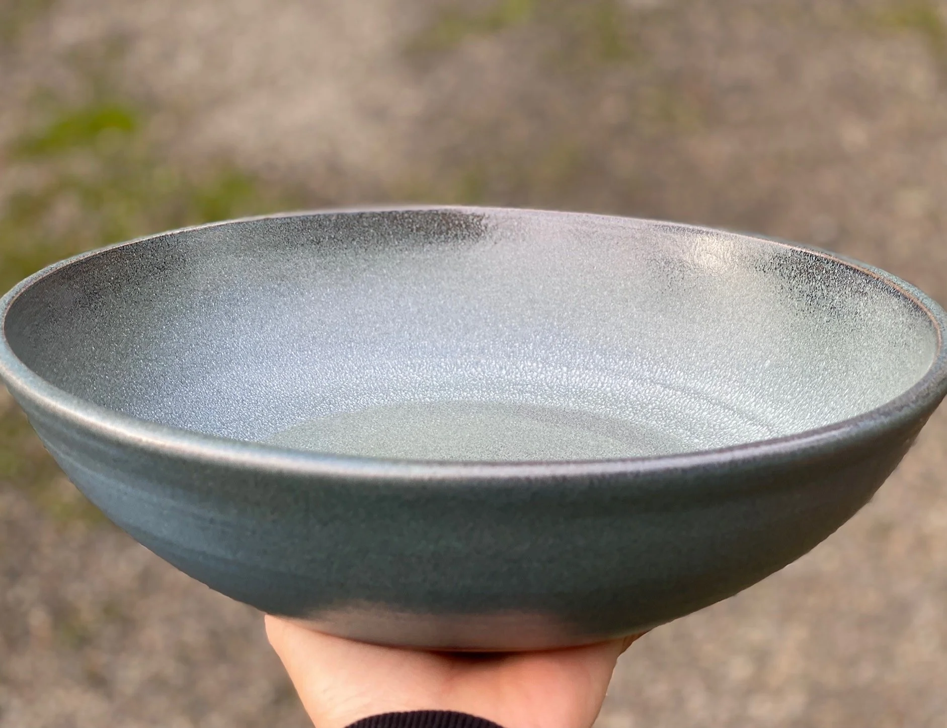 Hand holding a gray ceramic bowl with a matte finish outdoors with blurred ground background. Sonia Lesage Ceramics
