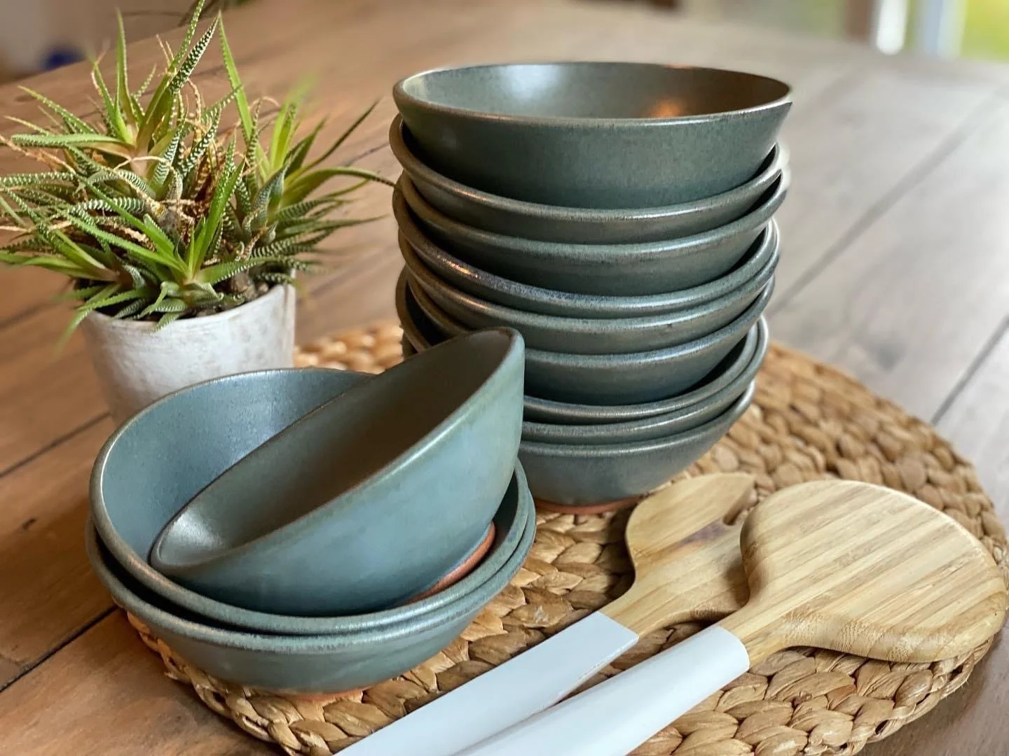 Stack of blue/grey bowls on woven placemat with a potted succulent plant and wooden spatula on wooden table. Sonia Lesage Ceramics