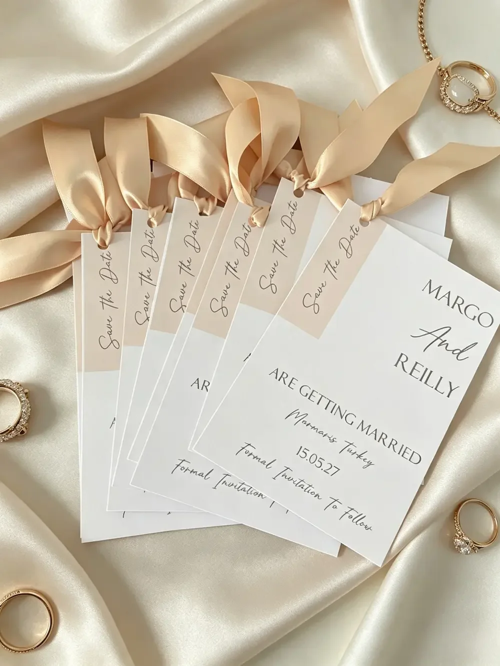 Elegant wedding save-the-date cards with beige ribbons, gold rings, and pearls, set on cream-colored satin fabric.