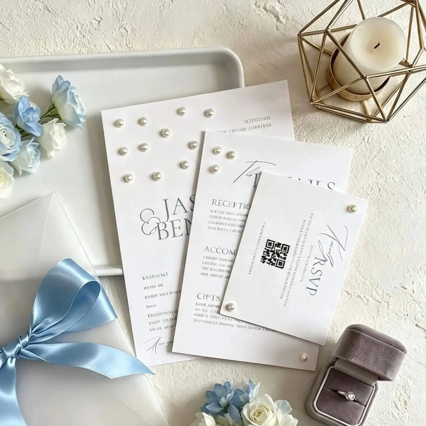Wedding invitation letters, a blue ribbon, a jewelry box with a ring, flowers, and a candle in a geometric holder on a white table.