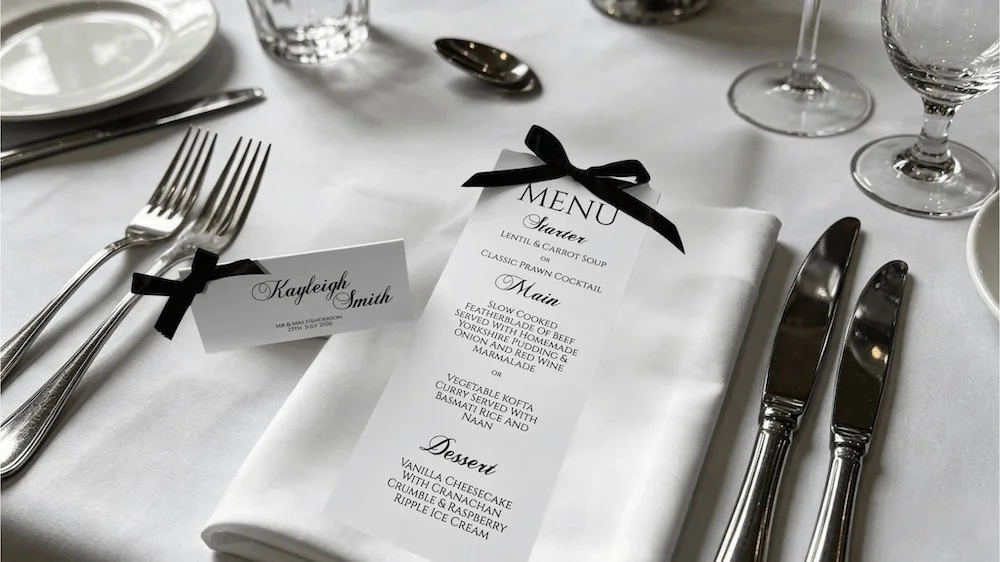 Formal dining setup with a white tablecloth, silverware, wine glasses, a place card, and a menu tied with a black ribbon.