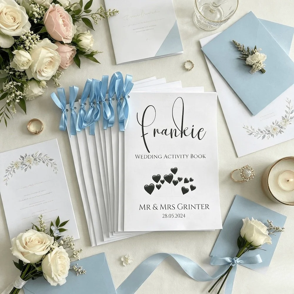 Wedding planning items including a wedding activity book for children, surrounded by floral arrangements, invitations, blue envelopes, a glass candle holder, a ring, and ribbons.